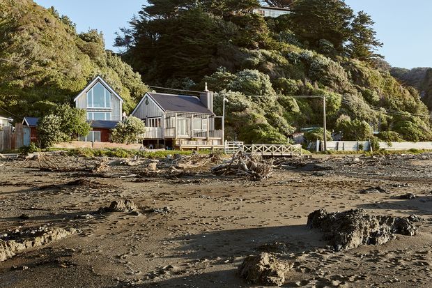 Exterior view of a group of compact coastal houses positioned directly behind a broad rocky and sandy foreshore. The buildings feature steep gable roofs, vertical weatherboard-style cladding, large glazed gable ends, and timber-framed windows and decks. Dense coastal vegetation and rugged hills rise behind the homes, while exposed mudflats and driftwood dominate the foreground.