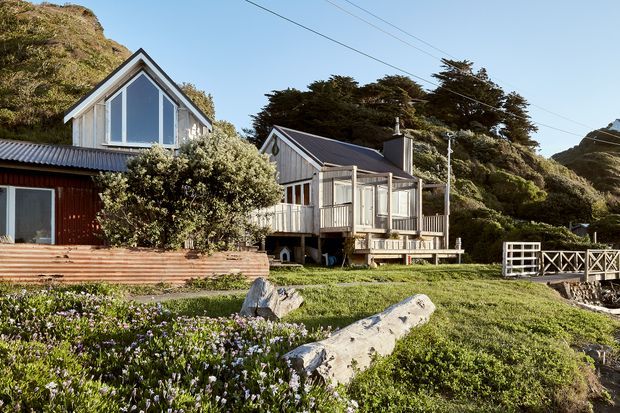 Exterior view of two small timber-clad coastal cottages on a grassy, rocky site. The buildings feature steep gabled roofs, large gable-end glazing, raised decks, corrugated metal roofing, and simple verandah structures. Dense coastal vegetation and exposed rock surround the homes, with a timber pedestrian bridge or deck railing at right and overhead utility lines visible.
