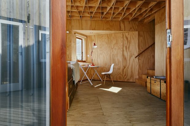 A compact timber-lined interior viewed through an open doorway, furnished as a small study or work area. Plywood walls, floor, built-in cabinetry and a distinctive exposed timber grid ceiling create a warm, utilitarian space. A small desk, white chair, task lamp, shelving and stair access are visible, with glazing at the sides.