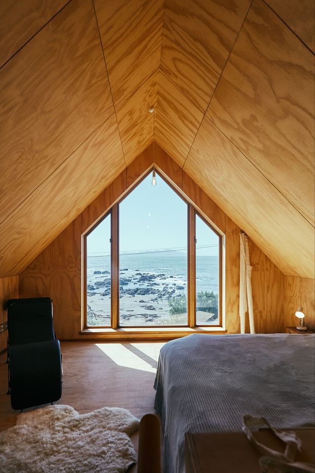 A warm, compact attic bedroom lined almost entirely in honey-toned plywood, with a steep vaulted A-frame ceiling and matching angled walls. A large triangular and vertical timber-framed window forms the focal point, offering a clear view of the rocky coastline and ocean. The room contains a bed with grey bedding, a black lounge chair, a small bedside lamp, and a soft cream rug. Natural sunlight creates a calm, intimate coastal retreat atmosphere.