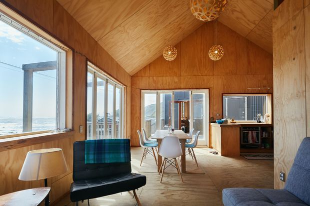 A bright wood-lined open-plan living and dining space with a sloped timber ceiling, large windows, and expansive coastal views. The room includes a black leather sofa, dining table with contemporary chairs, pendant lights, and a compact timber kitchen at the rear. Warm plywood surfaces and abundant glazing create a relaxed, natural atmosphere.