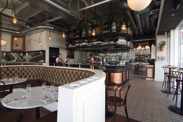 Interior view of a stylish restaurant or bar with an exposed industrial ceiling, extensive ductwork, hanging pendant lights, a central drinks bar with open shelving and plants, tufted leather banquette seating, marble-topped tables, timber and metal chairs, and patterned carpet flooring. The space combines industrial architectural elements with refined, classic hospitality furnishings.