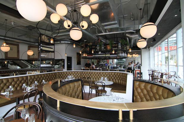 A stylish industrial-modern restaurant interior with curved, tan button-tufted banquette seating arranged around a central dining area. The space features black metal shelving, wine displays, exposed ceiling services and ductwork, globe pendant lighting, marble-topped tables, and large glazed windows along one side.