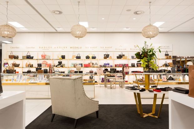 Bright, refined fashion and accessories retail interior with white display cabinetry and shelving, glass jewellery counters, handbags and shoes arranged throughout, and a central upholstered lounge chair on a dark rug. The space has a suspended white acoustic-tile ceiling with recessed lighting and decorative woven pendant lights, plus a display table with a tall floral arrangement.