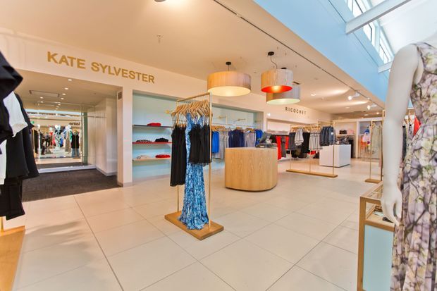 Bright contemporary fashion retail store interior with white tiled flooring, pale walls, extensive clothing displays, mannequins, timber display tables, suspended cylindrical pendant lights, and high clerestory glazing. Visible signage identifies Kate Sylvester and Ricochet retail areas.