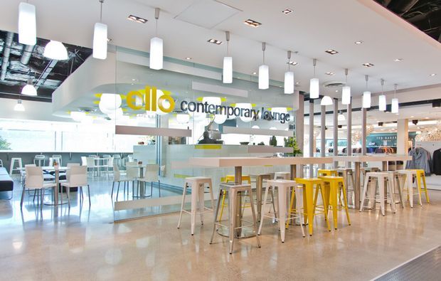 Bright contemporary commercial lounge or café interior in a shopping or hospitality setting. The open-plan space features a polished concrete floor, extensive glass partitions and counters, white pendant and recessed ceiling lighting, pale built-in surfaces, and high communal tables with mixed white, pale wood, and yellow stools. Exposed building services are visible in part of the ceiling, while retail activity is visible in the background.