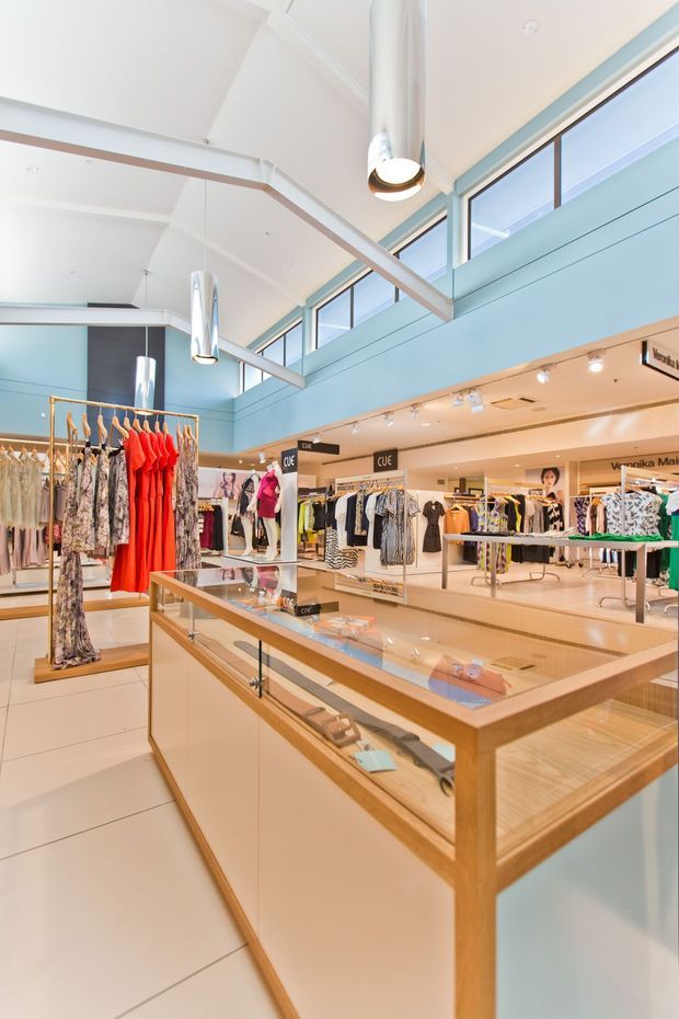 Bright, spacious multi-level retail clothing store interior with pale blue walls, high ceilings, clerestory glazing, and exposed structural beams. Clothing racks and mannequins surround timber-framed glass display counters and light-coloured tiled flooring. Pendant lights and track lighting illuminate the commercial fit-out.