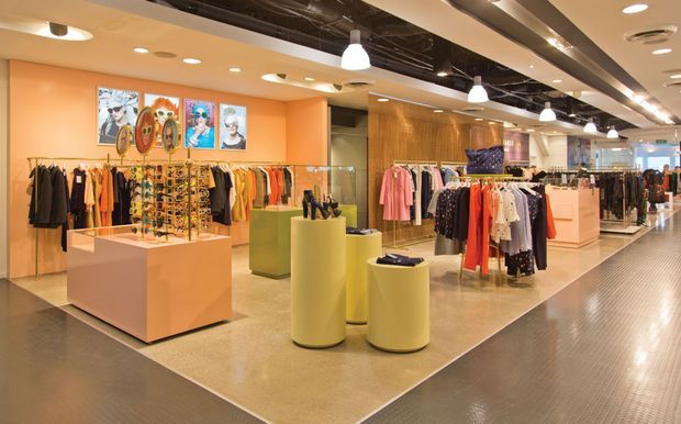 A brightly lit contemporary retail clothing store interior with warm peach walls, polished flooring, exposed dark ceiling services, recessed and pendant lighting, clothing racks, display tables, and cylindrical product plinths. Framed fashion artwork decorates the rear wall, while a wide aisle extends through the store toward the back.