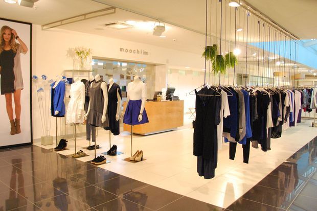 Bright contemporary retail clothing boutique with rows of garments on freestanding racks, mannequins, display shelving, a timber-toned checkout counter, hanging plants, polished tiled flooring, and extensive glass frontage. The space has warm white lighting and a clean, open layout.