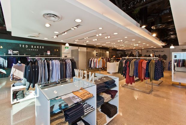 Interior view of a Ted Baker clothing retail store, with extensive garment racks, folded apparel displays, illuminated retail shelving, polished concrete flooring, and a contemporary suspended ceiling with recessed and track lighting. The store combines a bright white ceiling treatment with an exposed dark services ceiling toward the rear.