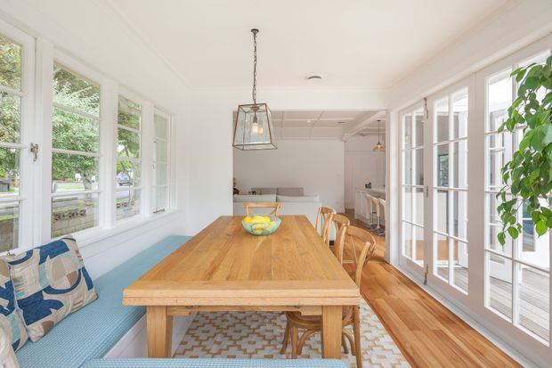 Bright, airy dining area in a glazed sunroom, featuring a large solid-wood dining table, built-in blue upholstered window seating, white-framed multi-pane windows and French doors, timber flooring, and a simple pendant light. The room opens toward a light-filled adjoining living or kitchen area and includes leafy outdoor views through the glazing.