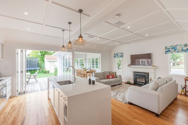 Bright open-plan kitchen and living room with a large white island, integrated sink and pendant lights in the foreground. The adjoining living area includes two light upholstered sofas, a fireplace with mantel and wall-mounted television, while a dining area and glazed doors open to a timber deck and garden. White coffered ceiling details and warm timber flooring create a fresh, welcoming interior.