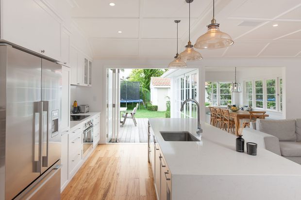 Bright open-plan kitchen and dining area with white shaker-style cabinetry, a large white island with integrated sink, stainless-steel refrigerator, pale timber flooring, and three glass pendant lights. The kitchen opens through glazed doors to a timber deck and garden, while a dining table and upholstered seating are visible to the right. The room has a light, refined, traditional coastal feel.