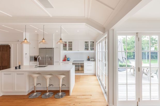 Bright, spacious white kitchen with a large island, three sculptural bar stools, stainless-steel appliances, glass-front cabinetry, pendant lighting, and warm timber flooring. The kitchen opens directly to a glazed outdoor deck or patio through full-height French doors, and features a decorative coffered ceiling with recessed lighting.