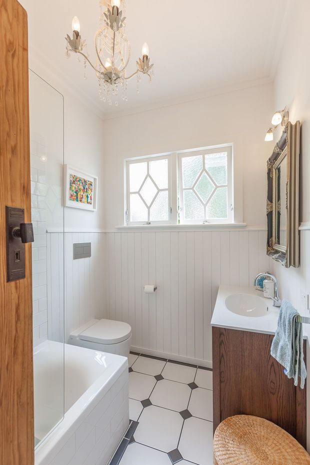 Bright, compact traditional bathroom viewed through an open timber door. The room contains a freestanding-style bathtub, toilet, timber vanity with white basin, ornate framed mirror, decorative wall lights, and a vintage crystal chandelier. White vertical wall panelling, white tiled surfaces, frosted geometric windows, and a white-and-dark geometric floor create a refined heritage-inspired interior.