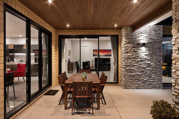 Covered outdoor dining terrace or courtyard adjoining a modern home, with a timber-lined ceiling, pale tiled floor, black-framed glazing, stacked-stone feature walls and a timber dining table with chairs. The glazing reveals a kitchen and living room beyond.
