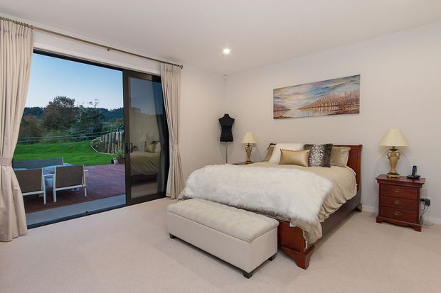 A bright, spacious bedroom with a large sliding glass door opening to a paved terrace and landscaped garden. The room features a wooden bed with layered neutral bedding, a long upholstered ottoman, matching bedside tables with lamps, full-height cream curtains, soft carpet, and a landscape artwork above the bed.