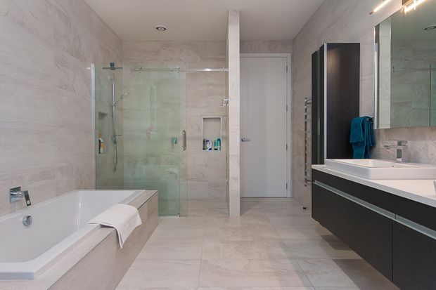 Contemporary, spacious bathroom with large-format pale stone-look wall and floor tiles, a freestanding-style bathtub, frameless glass shower enclosure, wall-hung dark vanity with a white countertop, large mirror, and recessed ceiling lighting. The room has a clean, refined neutral palette with strong contrast from the dark cabinetry.