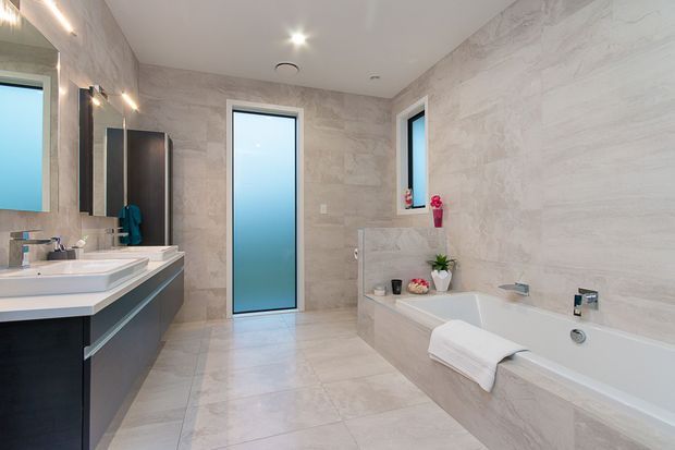 Contemporary bathroom with a freestanding-style built-in bathtub, double vanity, large-format pale stone-look wall and floor tiles, frosted glass doors and windows, recessed ceiling lights, and dark grey floating cabinetry. Pink floral accents and neatly folded towels add small decorative touches.