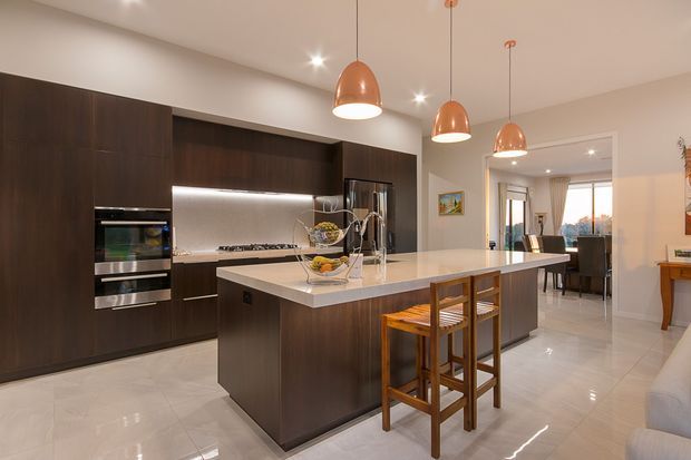 A spacious contemporary kitchen with dark timber-toned full-height cabinetry, integrated stainless-steel appliances, a large pale stone island with two timber bar stools, and a reflective light tiled floor. Three copper pendant lights hang above the island, while an open connection leads to a dining area beyond.