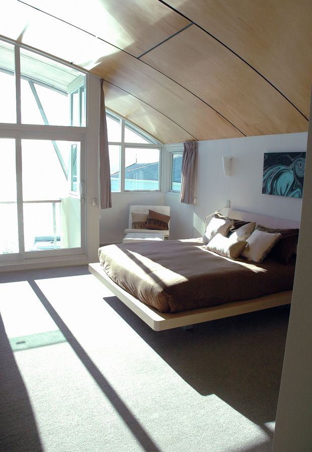 A bright, contemporary bedroom with a low platform bed, upholstered bedding, bedside lighting, and a lounge chair. Large windows and glazed doors provide abundant natural light and views outside, while the sloping timber-lined ceiling adds architectural character. The room has pale walls, neutral furnishings, and a soft carpeted floor.