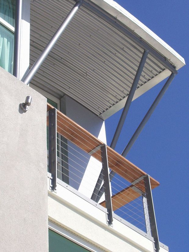 Upward-looking exterior detail of a contemporary building, showing a projecting corrugated metal canopy supported by slim dark steel posts, a balcony with timber-toned handrail caps and horizontal cable balustrading, smooth light-coloured rendered walls, and large glazing against a clear blue sky.