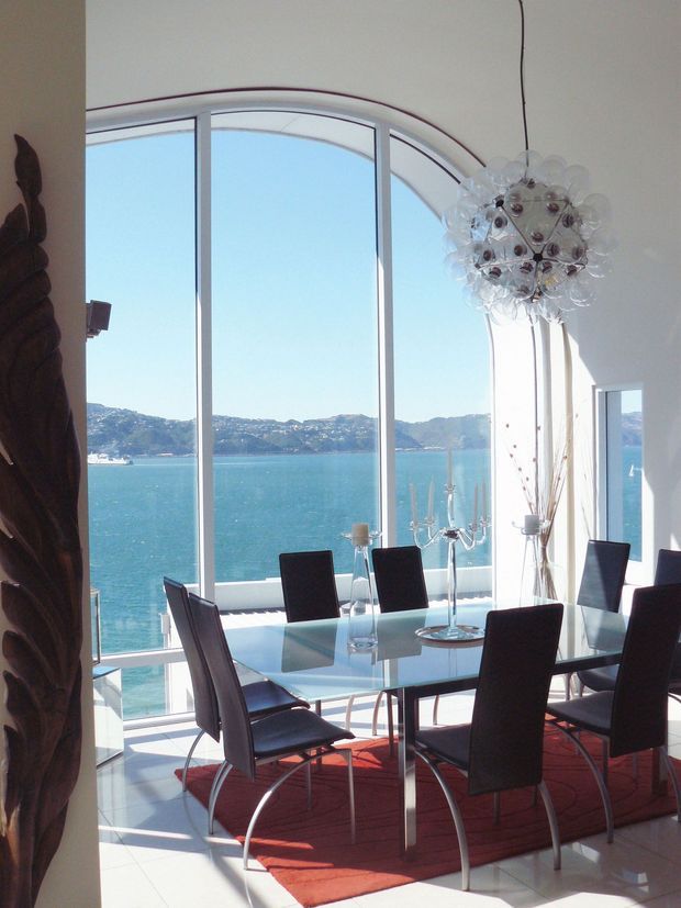 Bright waterfront dining room with a glass-topped rectangular dining table and dark upholstered chairs arranged beside expansive arched floor-to-ceiling glazing. The room has pale walls and tiled flooring, a red area rug, sculptural dark timber artwork, decorative candelabra, tall dried branches, and a spherical contemporary pendant light. A wide turquoise harbour and hilly shoreline are visible beyond the windows.