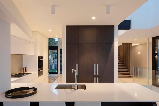 A bright, open-plan contemporary kitchen viewed from the island. The foreground features a large white island with an integrated sink, chrome tapware, and dark seating. White built-in cabinetry and appliances line the left side, while a tall dark timber pantry or storage wall forms the central feature. A glazed stairwell and timber-slatted staircase are visible beyond, with a double-height opening and glass balustrade contributing to the spacious, modern interior.