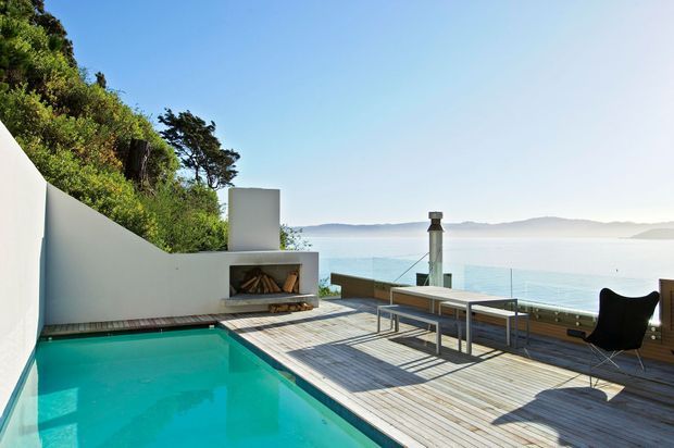 A modern waterfront terrace with a turquoise swimming pool, expansive timber decking, built-in outdoor fireplace, dining table, and lounge chair. Glass balustrades provide uninterrupted views across calm water toward distant hills, while dense vegetation rises behind a curved white wall.