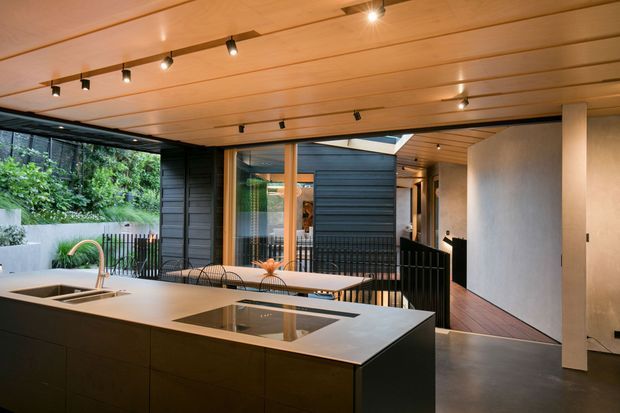 A contemporary open-plan kitchen opening onto a leafy outdoor area. The foreground features a large dark kitchen island with an integrated cooktop and a pale stone benchtop, while the rear has a dining table, dark horizontal cladding, extensive glazing, and a covered timber-lined ceiling with track lighting. A hallway and timber-floored interior space extend to the right.
