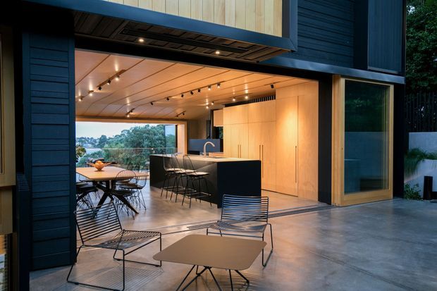 A contemporary open-plan kitchen and entertaining area with large retractable glazing opening onto an outdoor terrace. The space features a dark island with bar seating, pale timber cabinetry and ceiling panelling, polished concrete flooring, exposed dark exterior cladding, track and recessed lighting, and black wire-frame furniture. Trees and a distant landscape are visible beyond the opening.