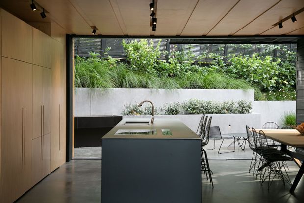 A sophisticated open-plan kitchen and dining area with a large dark island, integrated sink and sculptural tap, pale timber cabinetry, concrete-look flooring, and a warm timber-lined ceiling with track lighting. Full-width opening and glazing connect the interior to a planted courtyard with raised garden beds and a rendered boundary wall. A dining table with wire-frame chairs occupies the right side.