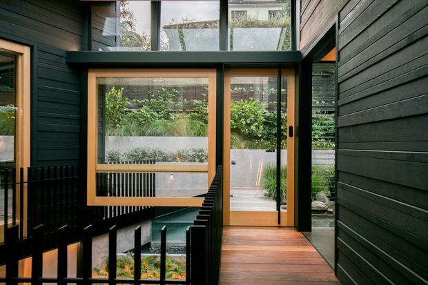A contemporary interior walkway or landing with dark horizontal timber wall cladding, warm timber-framed glazing, and a timber floor. Full-height windows and a glazed door open toward a planted courtyard, while a black metal balustrade borders a stair or lower-level void. High-level glazing brings in natural light and reveals neighbouring residential fabric beyond.