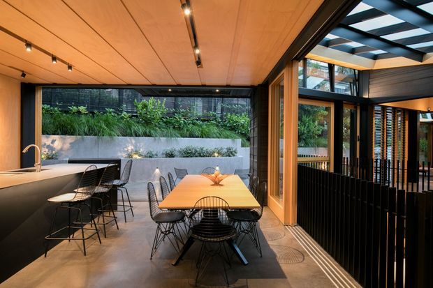 Contemporary open-plan dining and kitchen space with a long timber dining table, black wire-frame chairs, a dark kitchen island with brass tapware, and polished concrete flooring. Warm timber ceiling panels with integrated track lighting extend toward a glazed indoor-outdoor connection. A planted courtyard and black-framed glazing are visible beyond, with a covered exterior walkway or terrace to the right.