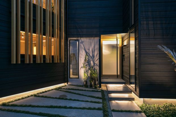 A dramatic contemporary residential entry courtyard at dusk, framed by dark horizontal cladding and tall vertical timber-like screening fins. Warm concealed lighting glows beneath the facade, through the screened windows, and along the stepping-stone path leading to a recessed front door. Gravel, planted joints, and ornamental grasses soften the minimalist architectural composition.