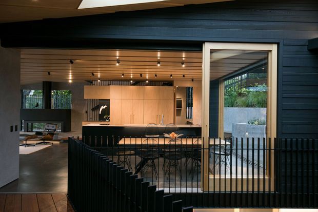 A dark, sophisticated contemporary kitchen and dining area viewed through black vertical metal balustrades. The space features extensive warm timber ceiling lining, dark horizontal exterior-style wall cladding, a pale timber kitchen with integrated cabinetry and island, black-framed glazing, and a dining table with wire-frame chairs. A living area is visible to the left, while landscaped greenery can be seen through the glazing.