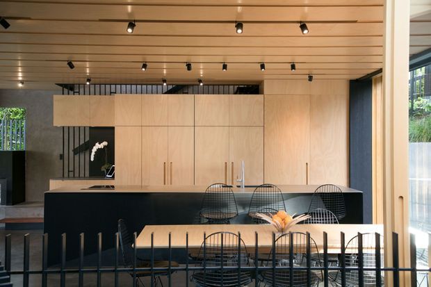 A contemporary open-plan kitchen and dining area with a large dark island, pale timber full-height cabinetry, integrated appliances, and a light timber slatted ceiling with recessed spotlights. A dining table with wire-frame chairs occupies the foreground, while expansive glazing and a timber-framed opening bring in daylight. The space has a clean, refined material palette with strong black-and-light-wood contrast.