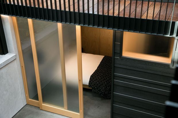 Compact loft-style sleeping nook viewed through partially open sliding doors, with a visible bed, warm timber framing, translucent panels, dark horizontal wall cladding, and a black metal mezzanine guardrail above. The space has a contemporary, compact architectural character.
