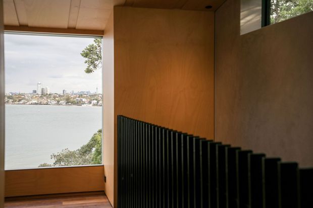 Interior architectural view of a compact stairway descending beside warm timber wall and ceiling linings. A black vertical slatted balustrade forms a strong diagonal in the foreground, while a large window frames a waterfront and urban skyline beyond. The space has a restrained, contemporary character with strong natural light and water views.