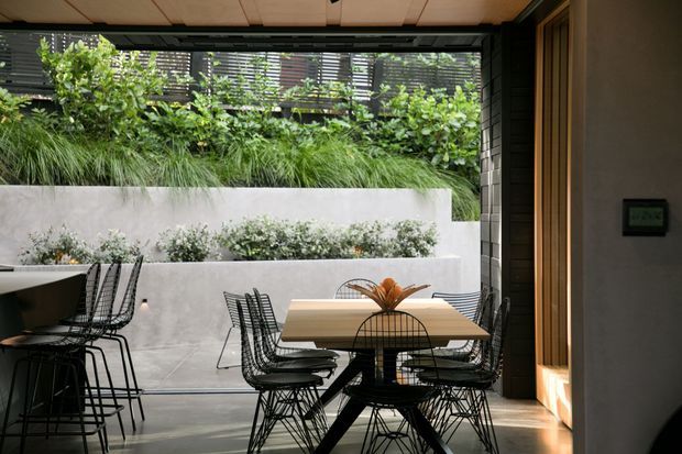 A contemporary indoor-outdoor dining area with a large open glazed wall overlooking layered planter beds and greenery. The space contains a long light-wood dining table, black wire-frame dining chairs, a black counter with stools at left, dark tiled wall surfaces, warm timber ceiling panels, and timber-framed glazing. Natural light and the planted courtyard create an airy, refined atmosphere.