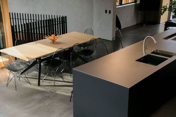 Open-plan contemporary kitchen and dining area with a large dark island, integrated double sink and curved metal tap. A light timber dining table is surrounded by black wire-frame chairs, with a black vertical screen feature against a textured light wall. Concrete flooring and dark cabinetry create a restrained industrial-modern feel.