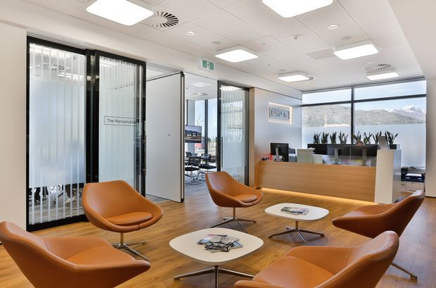 Bright contemporary office reception and waiting area with warm timber flooring, white acoustic ceiling tiles, black-framed glazed partitions, frosted glass doors, a timber reception desk, and several caramel leather lounge chairs around low white coffee tables. Large windows provide views of a mountain landscape, while indoor plants and framed artwork add finishing touches.
