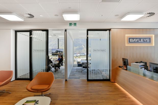 A bright contemporary commercial reception area with a timber-look floor, pale walls, acoustic tile ceiling and recessed lighting. A timber-paneled reception desk and illuminated Bayleys sign occupy the right side, while glazed black-framed office partitions and doors reveal workstations beyond. Orange lounge chairs add a warm accent, and mountain scenery is visible through the rear glazing.