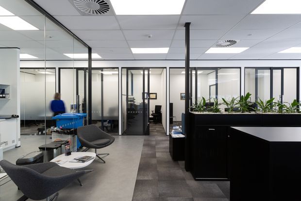 Contemporary commercial office interior with a reception counter in the foreground, black-framed glazed meeting rooms and offices, a waiting area with modern lounge chairs, and indoor planting. The space has a suspended acoustic ceiling with recessed fluorescent lighting and exposed air vents, polished concrete-look flooring in the lounge area, and dark modular carpet tiles through the circulation zone.