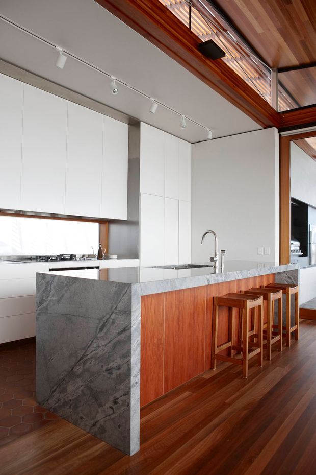 Bright contemporary kitchen dominated by a large island with a grey veined stone waterfall benchtop and warm timber cabinetry. White full-height cabinetry lines the rear wall, with a cooktop and sink visible. Timber flooring, exposed timber ceiling elements, clerestory glazing, and track lighting create a clean but warm interior. Timber bar stools sit along the island.