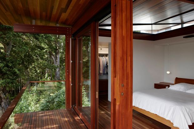 A timber-lined bedroom opening onto a forested balcony through extensive glazed doors. The room features rich reddish timber framing and flooring, a white bed with minimal built-in bedside furniture, and a partially visible wardrobe. A glazed roof or clerestory-like section brings natural light into the bedroom, while the balcony and surrounding trees create a strong connection to nature.