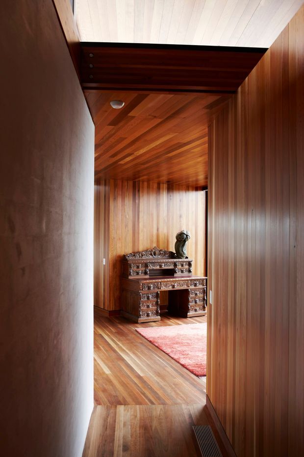 A narrow, richly timber-lined interior passage opens into a small formal room or study. The walls, ceiling, and floor are clad in warm medium-to-dark wood, with a carved antique-style desk or console, decorative sculpture, and a red patterned rug. A white painted wall and ceiling section at the foreground contrast with the extensive timber panelling. The image has a warm, refined, heritage-inspired character.