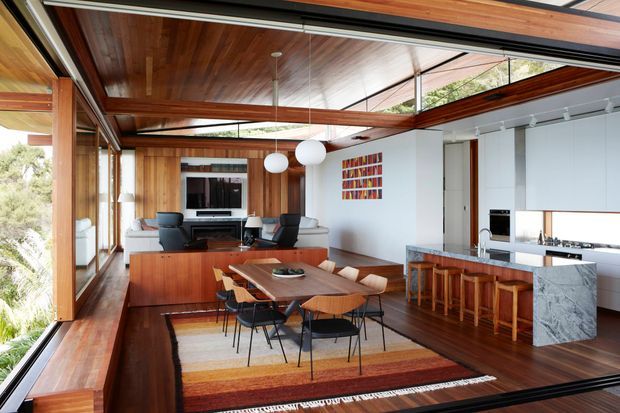 Open-plan contemporary living, dining and kitchen interior with extensive floor-to-ceiling glazing, warm timber ceilings and floors, a timber dining table, built-in media joinery, and a marble-clad kitchen island. Exposed beams and layered timber roof planes create a strong connection between the interior and the surrounding landscape.