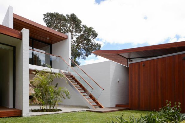 Exterior view of a contemporary residence organized around a landscaped courtyard. White rendered walls, warm timber soffits and cladding, broad glazing, and an exposed external stair with slim metal handrails create a clean modern composition. A separate timber-clad volume with a projecting roof is visible to the right, with lawn and planting in the foreground.