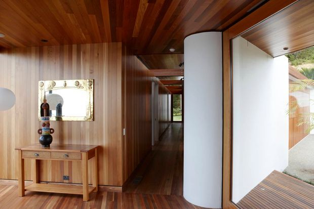 A warm, timber-lined interior hallway or entry passage with rich wood flooring, vertical wood wall panelling, and a matching timber ceiling. A narrow console table with a decorative mirror and vase sits along the left wall. The passage leads toward a glazed opening and an exterior timber deck, while a curved white partition creates a striking contrast against the dark timber finishes. The image has a refined mid-century-inspired architectural character with strong indoor-outdoor connection.